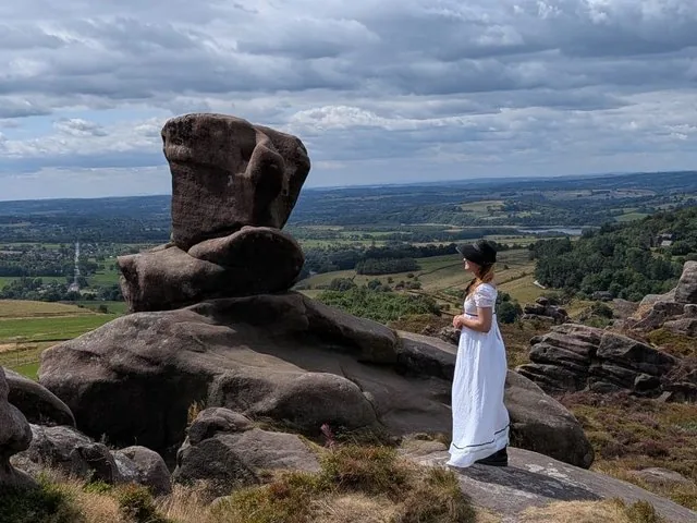 Una persona con un vestido blanco de pie sobre unas rocas mirando un paisaje.