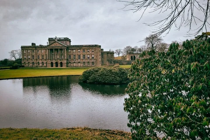 Ein großes Herrenhaus, betrachtet über einen Teich mit einer grünen Rasenfläche.